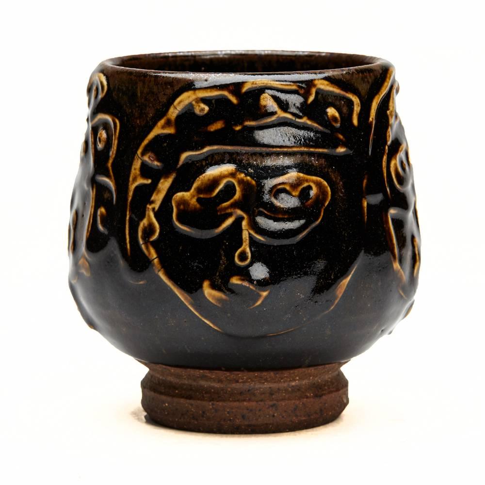 A stunning vintage American Studio Pottery tea bowl decorated with Expressionist faces by Peter Voulkos. The stoneware tea bowl stands on a narrow rounded unglazed foot with a rounded cup shaped body decorated in dark tenmoku glazes with relief