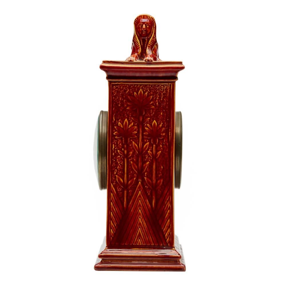 A rare Burmantofts Faience mantel clock of tall tapering rectangular form, cast in low relief with a heron wading in a river, with panels of tall flowering stems, the top surmounted with a Sphinx. The clock is glazed in red. The clock is model