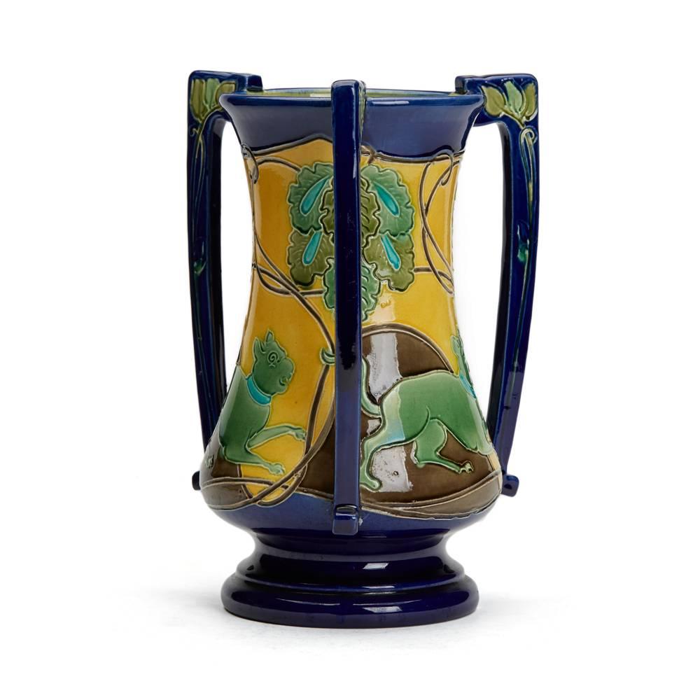 An unusual and rare Art Nouveau Burmantofts Faience tyg vase of waisted cylindrical form with three applied handles, incised with dogs running between foliage on sinuous Art Nouveau stems. The vase is painted in green, brown, blue and turquoise on a
