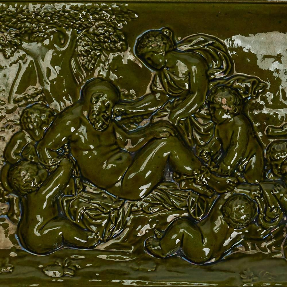 Scarsa piastrella baccanale di Burmantofts Faience di forma rettangolare modellata in rilievo con figure che assistono un uomo che riposa all'interno di un bordo a gradini rialzato e decorato con smalti verdi. Marchi incisi di Burmantofts, marchio