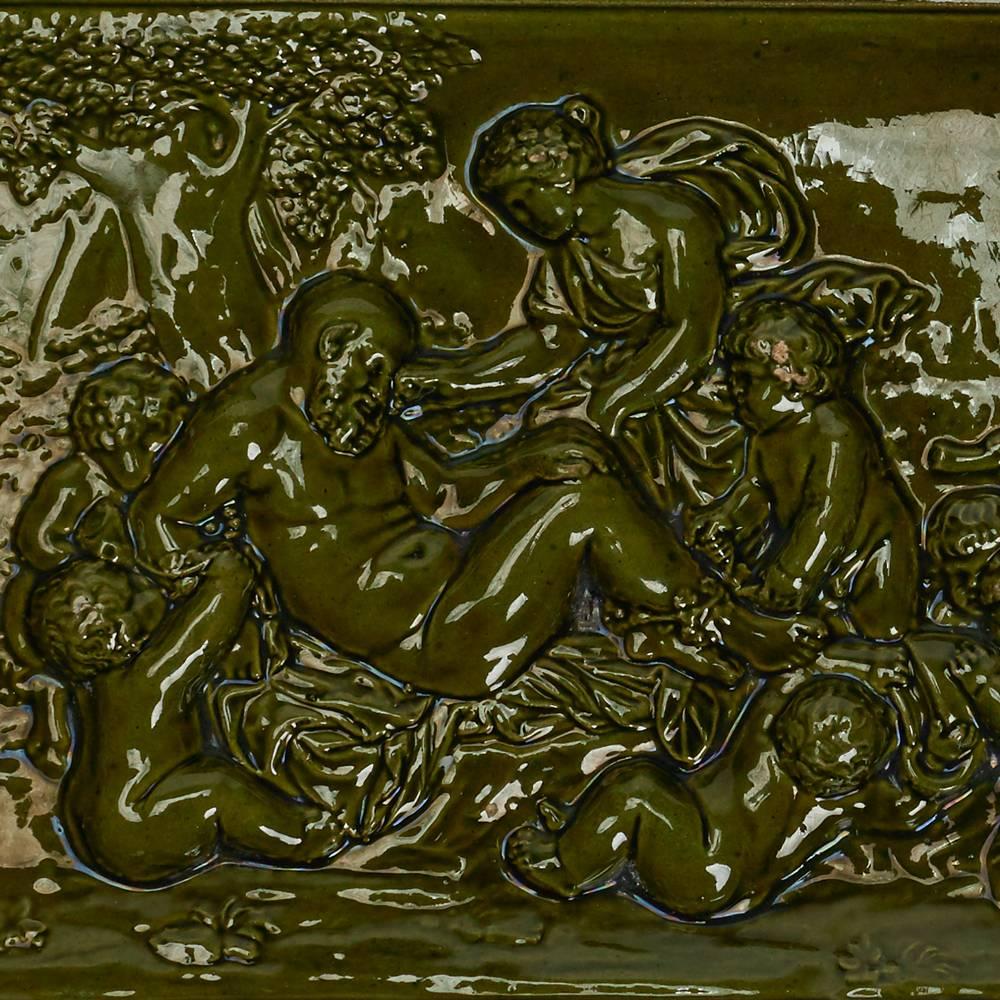 Rare carreau bacchanalien en faïence de Burmantofts, de forme rectangulaire, modelé en relief avec des personnages s'occupant d'un homme au repos, dans un cadre en gradins et décoré de glaçures vertes. Marques Burmantofts incisées ainsi que la