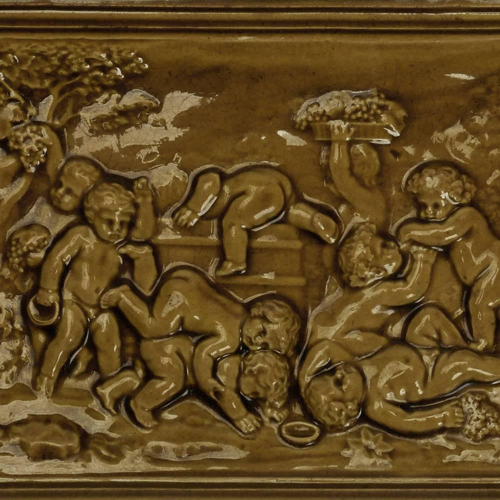 Rare carreau bacchanalien en faïence de Burmantofts, de forme rectangulaire, modelé en relief avec des personnages cueillant des raisins, certains jouant, dans un cadre surélevé à bord étagé et décoré de glaçures vert moutarde. Marque incisée à la