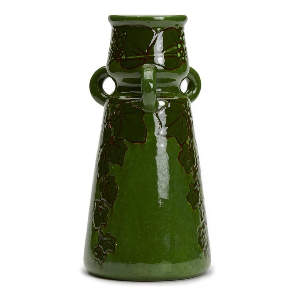 An unusual Burmantofts Faience vase of waisted tapering cylindrical form with three loop handles and with trailing ivy to the body, the neck with a similar band with fruiting ivy, painted in dark green on a light green ground. The vase has impressed