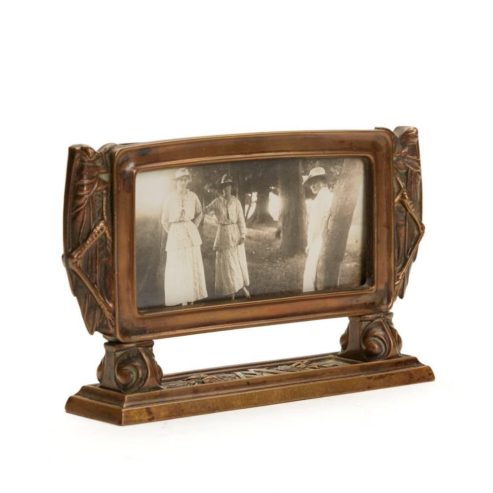 An unusual and stylish Arts & Crafts bronze frame mounted on a long rectangular base with moulded grasshoppers perched on either side of the frame. The rectangular shaped frame has an inserted glass and has the initials MV relief moulded to the