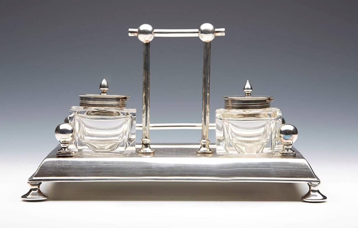 A stylish Arts & Crafts silver-plated desk stand comprising of two glass lidded inkwells together with a pen recess. The set is of rectangular shaped raised on four rounded feet with a raised central handle and surround with ball terminals. The