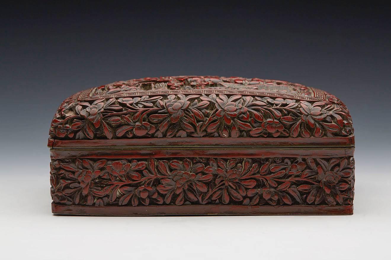 A fine antique Chinese Qing red cinnabar lacquered lidded box with a relief carved central village scene with figures set within a floral surround. The metal cased box with removable cover and black lacquered base.