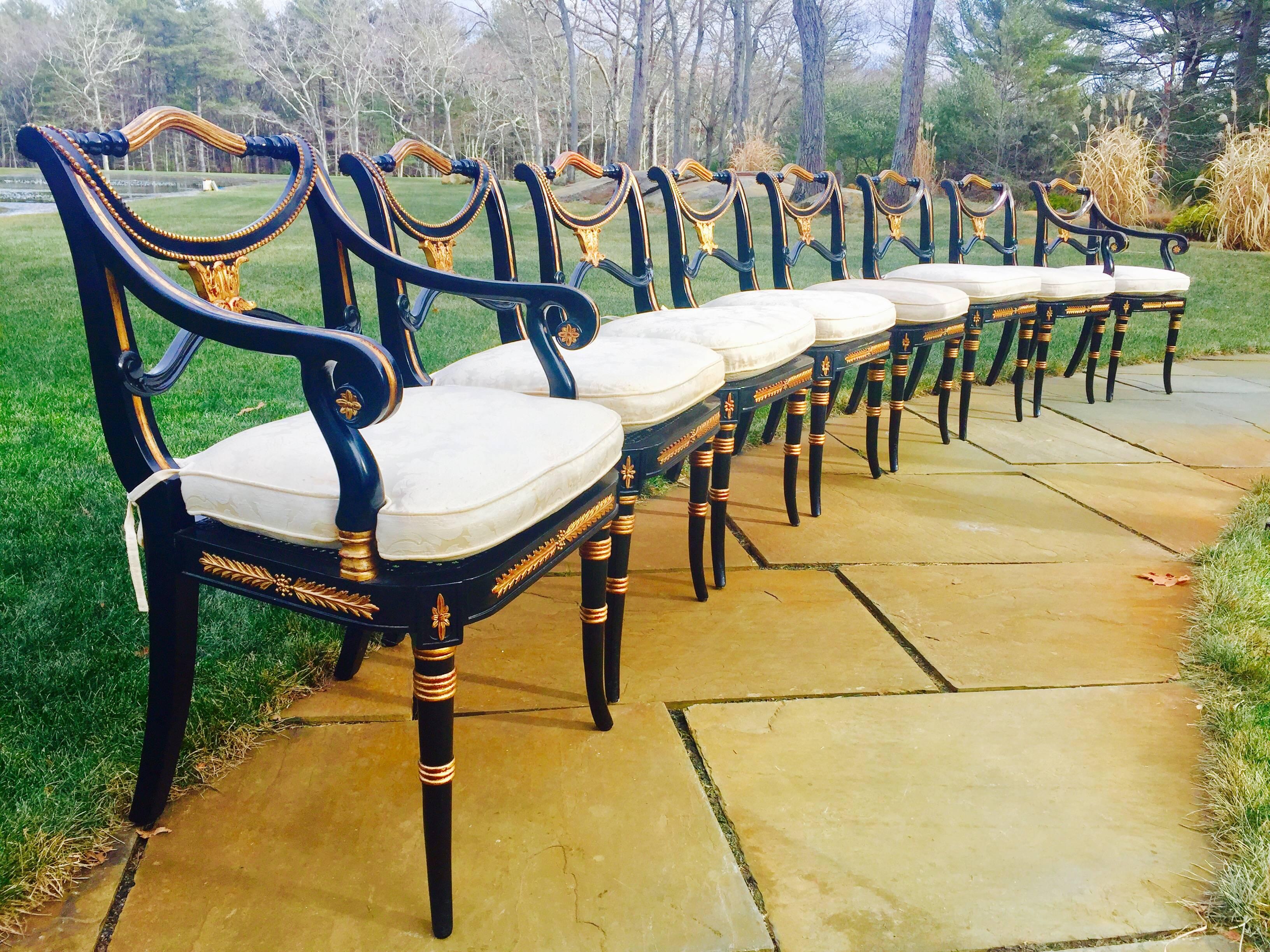 This Classic set of eight Regency style dining chairs was produced by Theodore Alexander. 
It is hand-carved of mahogany and has been ebonized and gilded to highest standards. The seats are caned and have custom cushions on top for ultimate comfort.