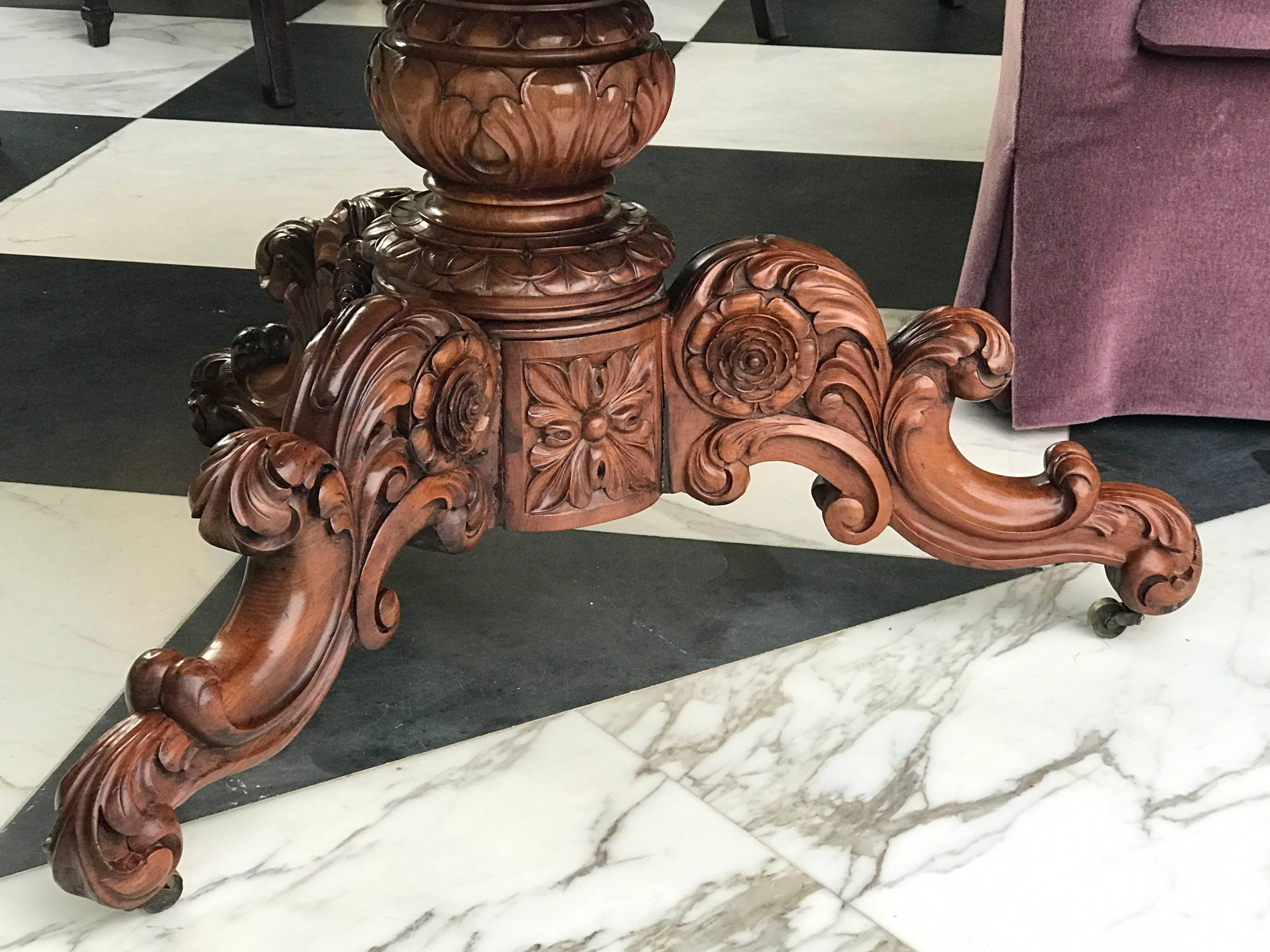 This elaborate mahogany center table was manufactured in England in the Regency period. The base is heavily carved in a foliate design incorporating three dimensional roses and rests on its original brass castors. The rich rouge marble top is a
