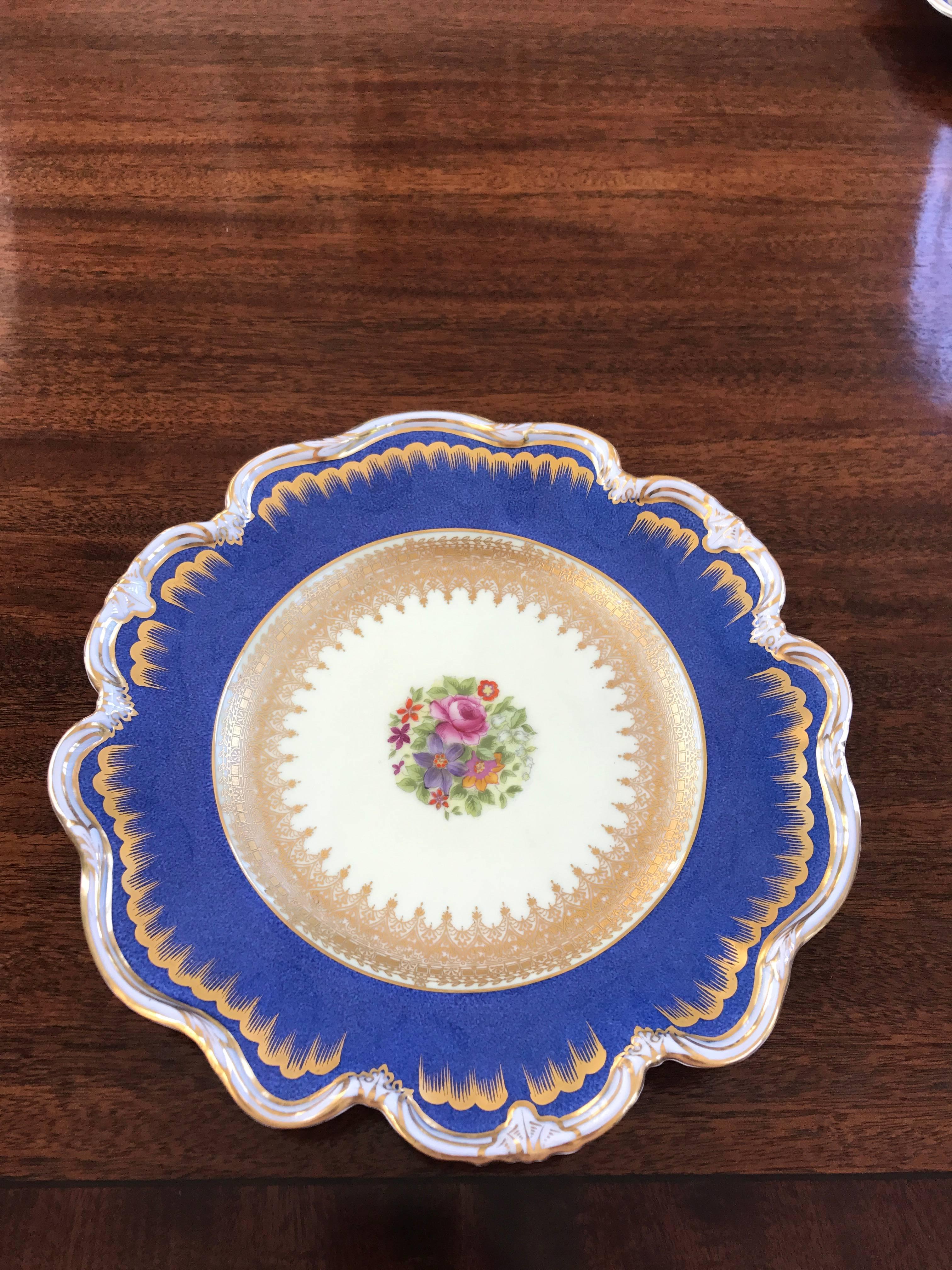 A lovely set of ten scalloped porcelain plates, each with a 24-karat gold trim and a broad cobalt blue border. 
The center is adorned with a hand painted bouqet of flowers on an ivory background. 
Made in England
George Jones & Sons.
Circa 1900

