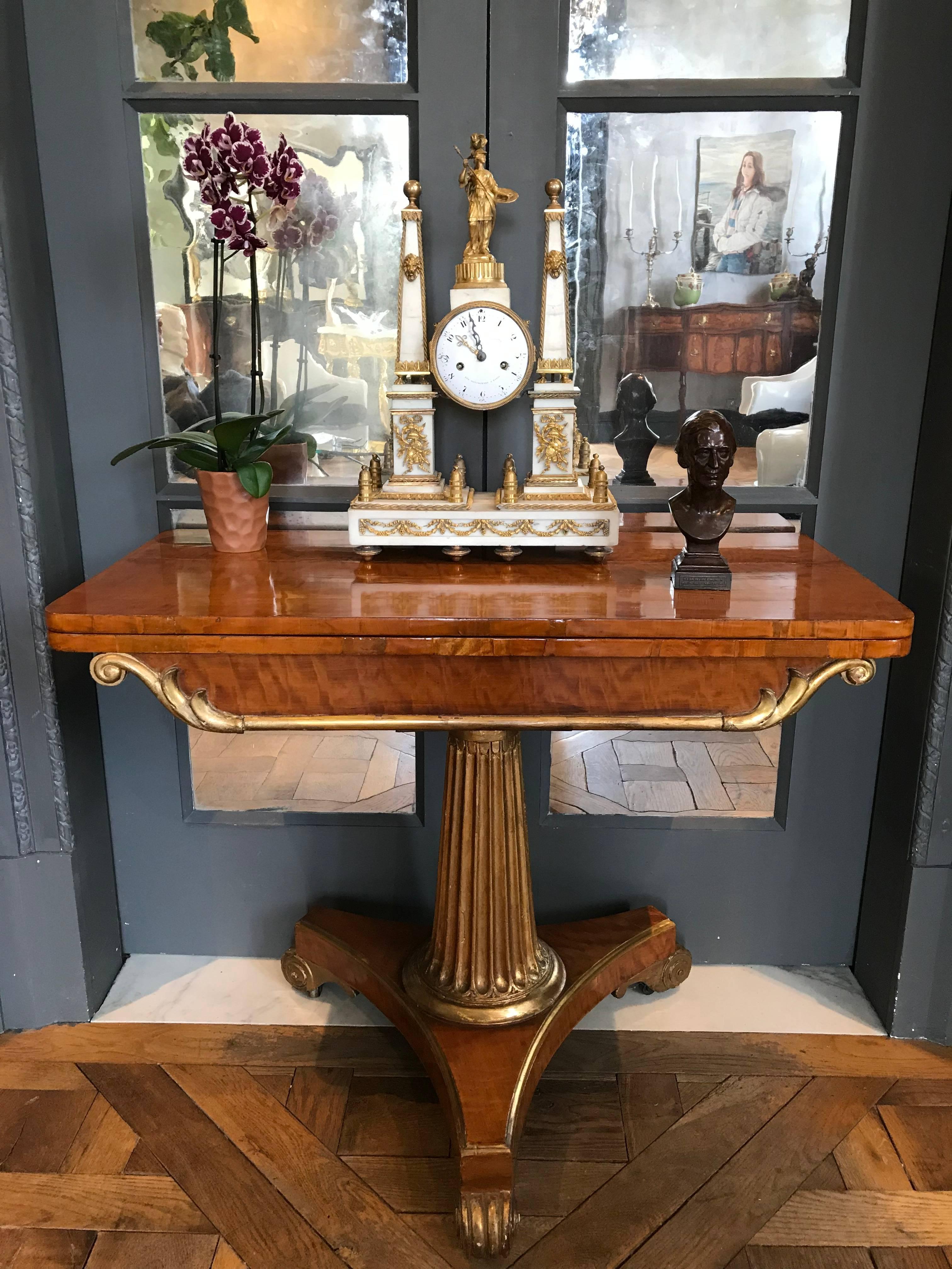 This elegant English neoclassical period card table is veneered in flame birch and has gilded elements. Three gilded scroll feet support the Regency trefoil base out of which a graduated reeded column rises. The top swivels and reveals the storage