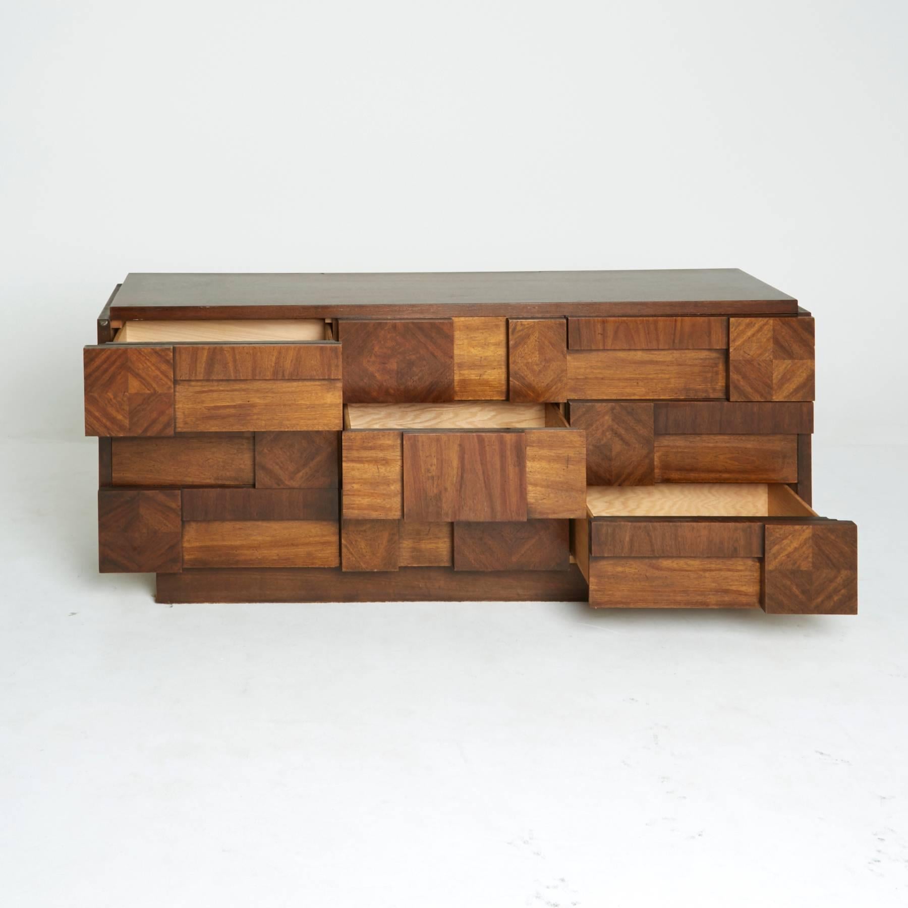 This Brutalist credenza, created by Lane, features a cubist style wood arrangement across the front of the nine dresser drawers. Each of the blocks of wood has a rich and unique grain, each going a different direction to create a collage of