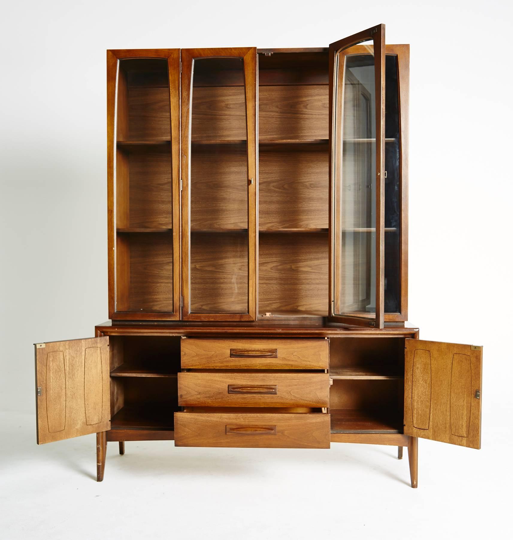 This large American modern hutch unit has beautiful Midcentury details adorning its exterior and ample storage inside. The top china cabinet has four windows that are tapered at the top and the bottom, two of which are doors that open into the