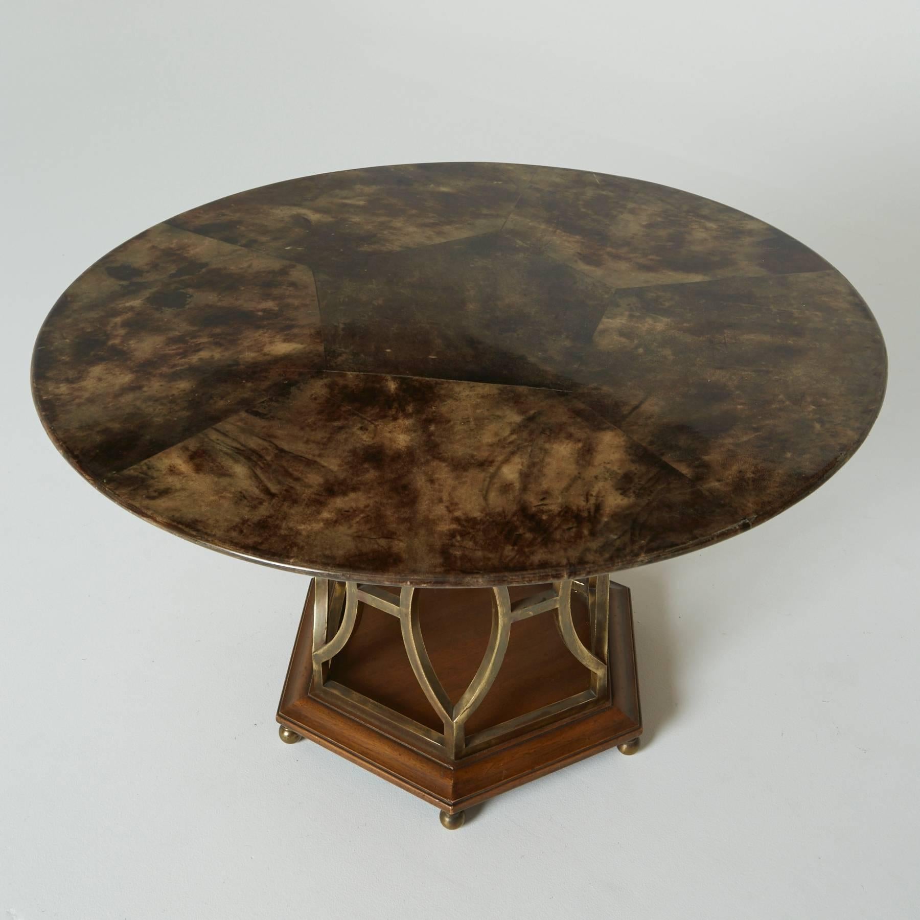 Brown lacquered goatskin table top is marbled in texture, with a polygonal pattern which is mirrored throughout the whole piece. The round dining surface rests on angular wooden mounts, below, a sculptural brass table base which is rich with
