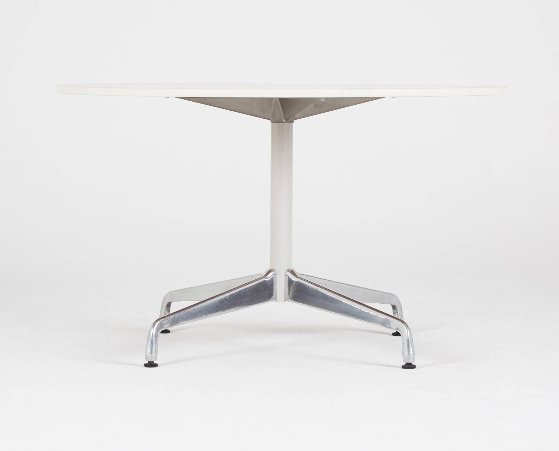 A 1964 design by Ray and Charles Eames for Herman Miller. A sleek, sophisticated dining table with a round white laminate top, perched on a cylindrical base with four (4) curved aluminum legs and black sliders. Gather your people together around