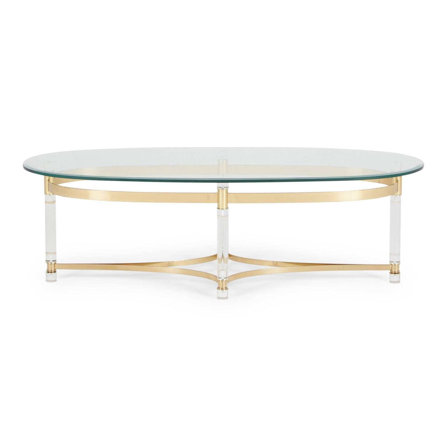 Elegant and wonderfully chic Lucite and brass coffee table in the style of Charles Hollis Jones and Maison Jansen. This oval-shaped piece is comprised of a glass table surface atop a brass frame with lucite legs. The Regency style, with the