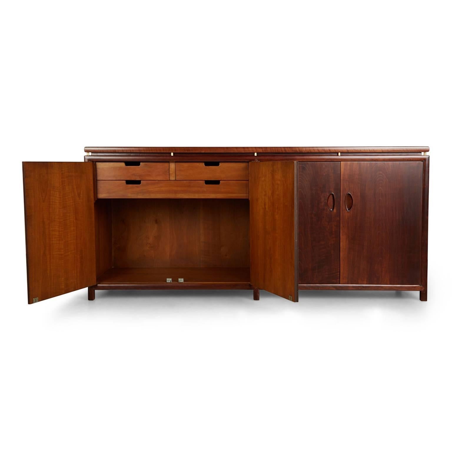 This circa 1960s restored Michael Taylor for Baker credenza / buffet comprised of solid walnut with stunning wood grain, cylinder shaped brass spacers that connect the top to the cabinet and bares the manufacturers stamp inside one drawer. The piece