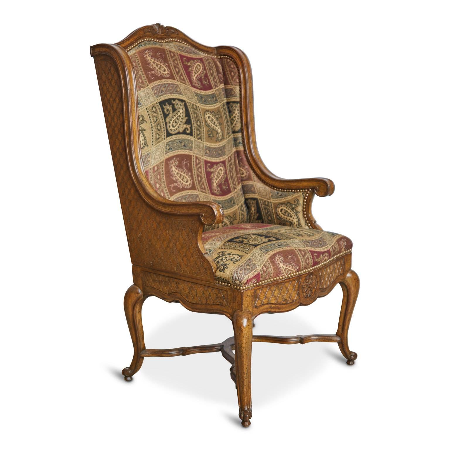 Louis XVI style wingback chair comprised of a carved oak frame and paisley upholstered seats. The curvaceous frame of this royal looking armchair features scrolled arms, cabriole legs with an adjoining X-base, feuilles d'acanthe sculpted on the