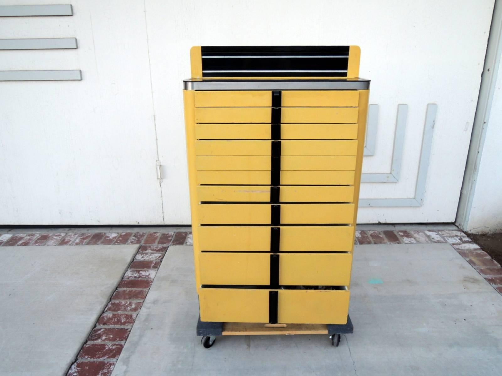 Fantastic Art Deco dental cabinet made by the American Cabinet Company, USA. There are black plastic handles and a painted yellow enamel frame and drawers. There are ten drawers total and the fourth drawer down is a double drawer. The top area of