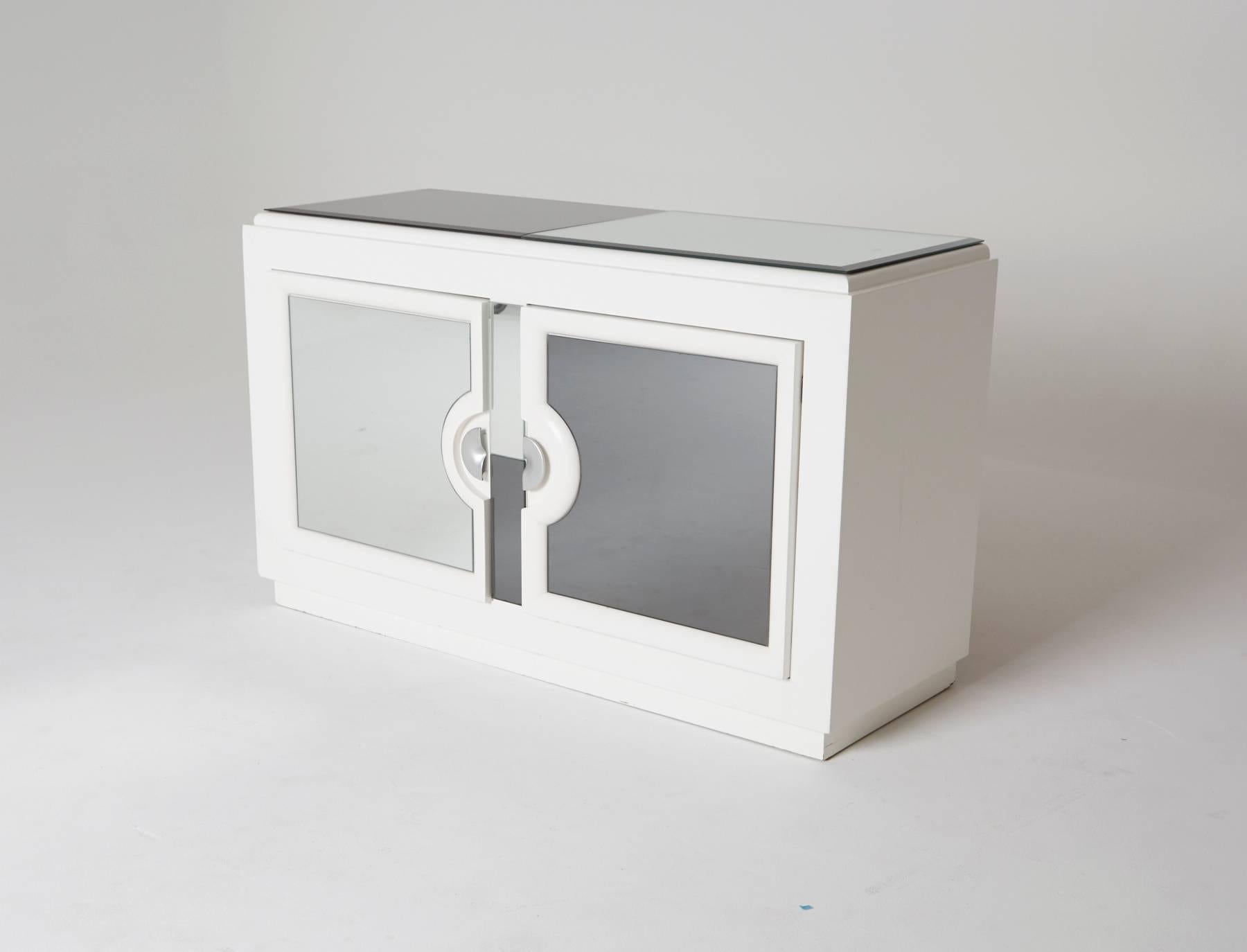 This Paul Laszlo styled credenza has a clean white lacquered frame and glass surfaces, alternating between mirrored and smoked, on the front face and the top. The cabinet doors, with adjoining half-circle handles, open two storage compartments; One