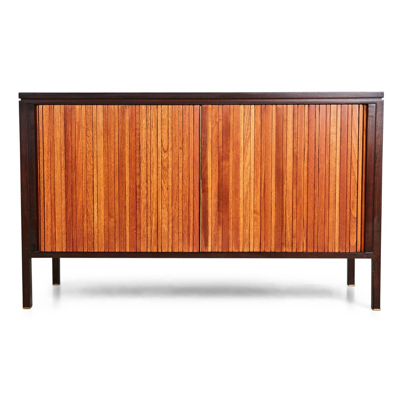 Designed by Edward Wormley for Dunbar, this innovative, inspired piece functions as both a credenza and a pop-up table, enabling it to operate in numerous settings. Comprising of teak tambour doors with brass lip hardware, which slide and retract