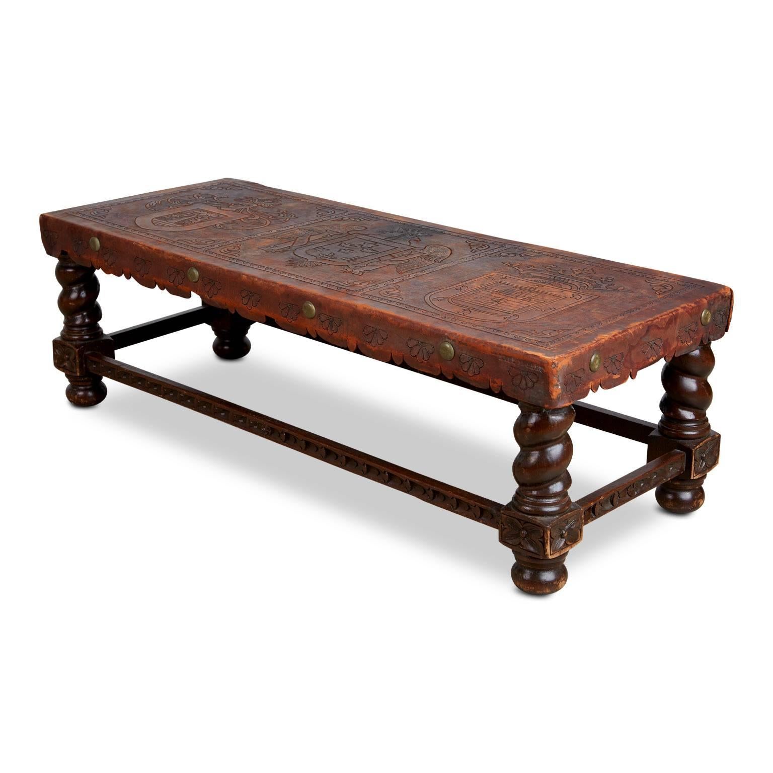 Beautiful hand tooled Peruvian piece which can be either used as a bench or coffee table. Comprised of turned wood legs with a dark stain and leather top illustrated with embossed shields. The sides of this piece feature scalloped details with