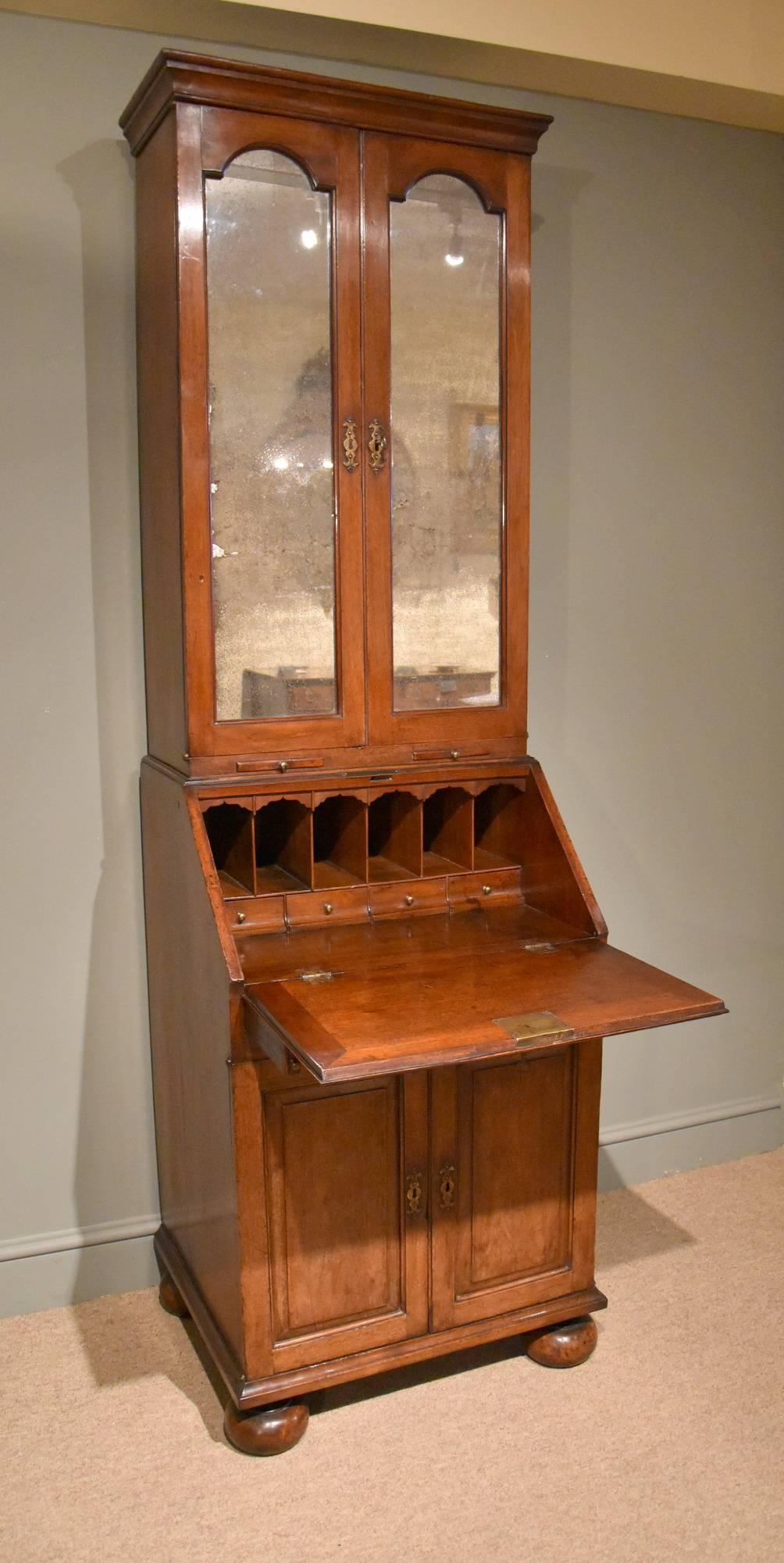 A rare and unusual early 18th century solid walnut bureau bookcase of small size. Mirrored doors enclosing adjustable shelves below cupboard doors. The bureau with pigeon holes and an arrangement of four drawers is enclosed by writing tall. The