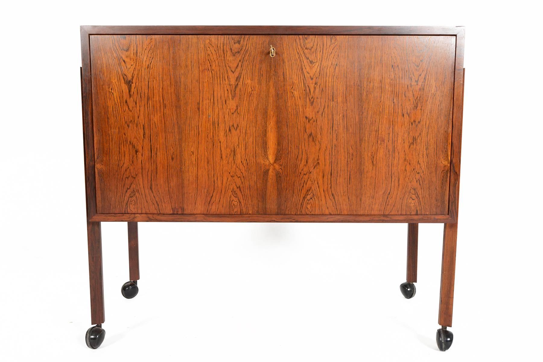 This Danish modern Mid-Century bar cart in Brazilian rosewood will be a wonderful addition to any modern home. This wonderful cart features a large drop down door which opens to expose an oak lined interior with six laminate lined bottle holders.
