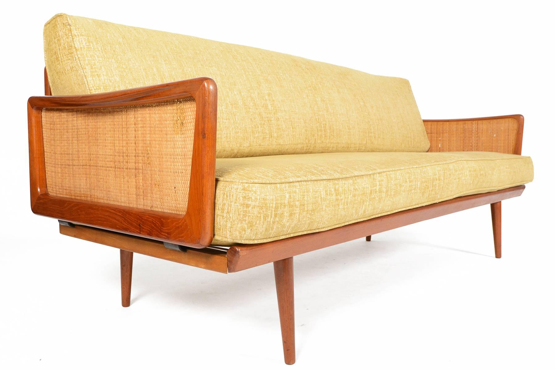 This fantastic Danish modern sofa was designed by Peter Hvidt in the 1960s. The solid teak frame features original cane arm panels inside the sculpted teak armrests and back paneling. Adorned with brass hardware. The two cushions are covered in a