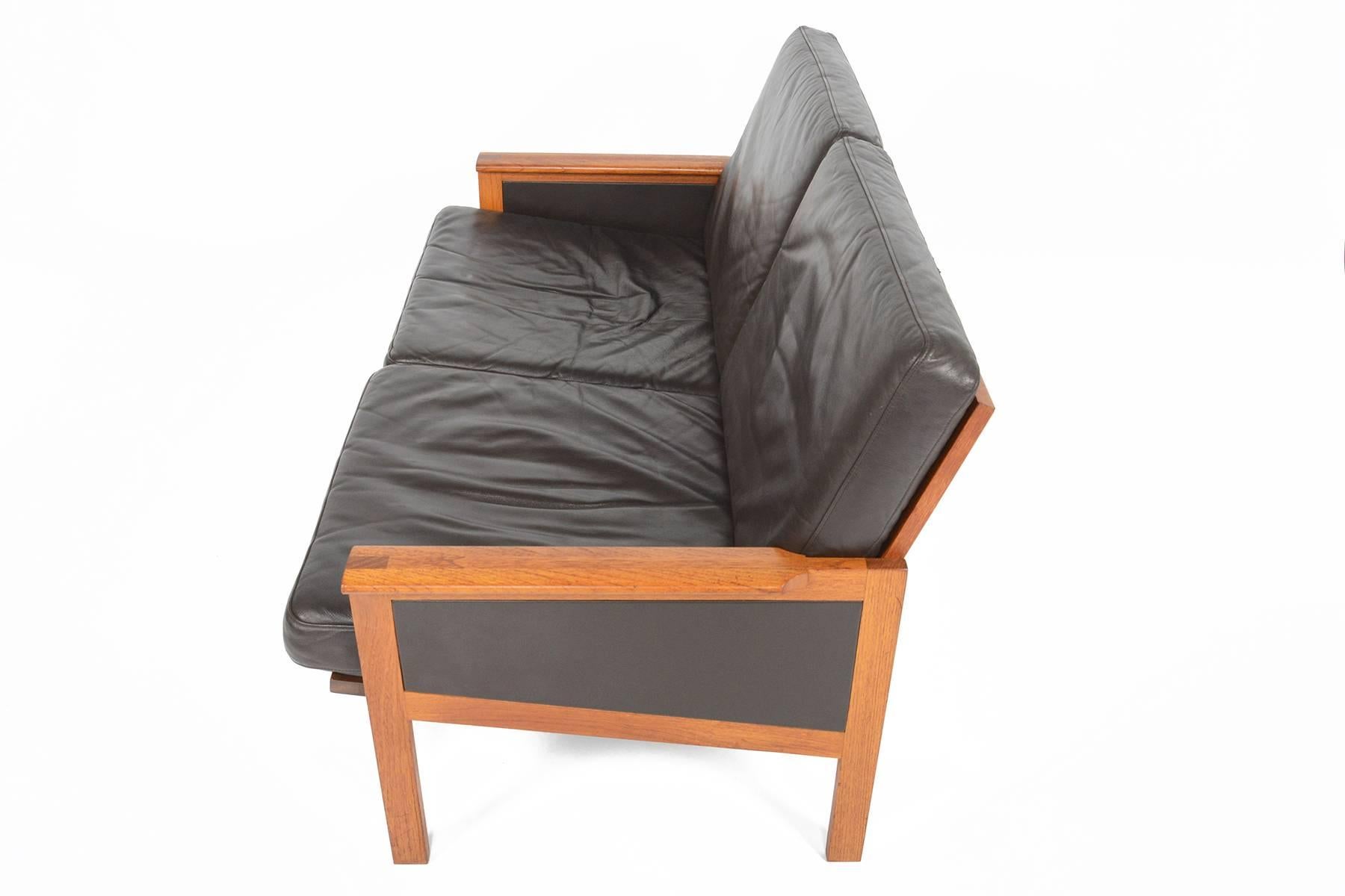 This beautifully sculpted teak loveseat sofa known as the ' Capella' model was designed by Illum Wikkelsø for Niels Eilersen in 1959. Expertly crafted from solid teak, this sofa features leather panels underneath each armrest for a truly modern