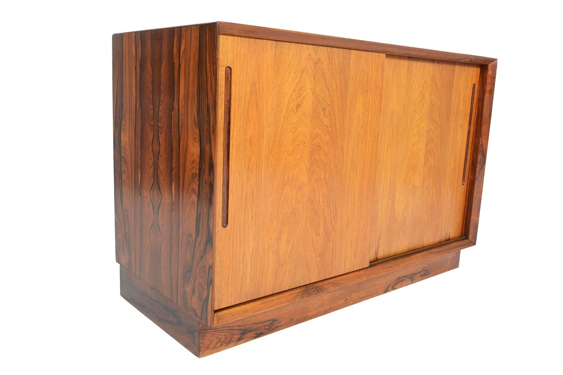 This Danish modern Mid-Century medium sized rosewood credenza offers two sliding doors accented by carved pulls. Doors open to reveal open storage with a removable shelf. Case sits on a solid plinth base completing its elegant Silhouette. In
