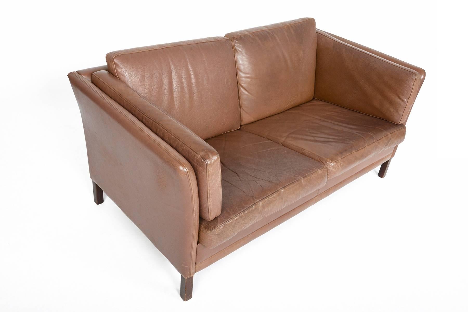 This beautifully sculpted loveseat sofa was manufactured by Stouby in the 1960s. This simple flared arm design offers an elegant profile. Six patinated leather cushions nest in the fully upholstered frame. Finished in square tapered mahogany legs.