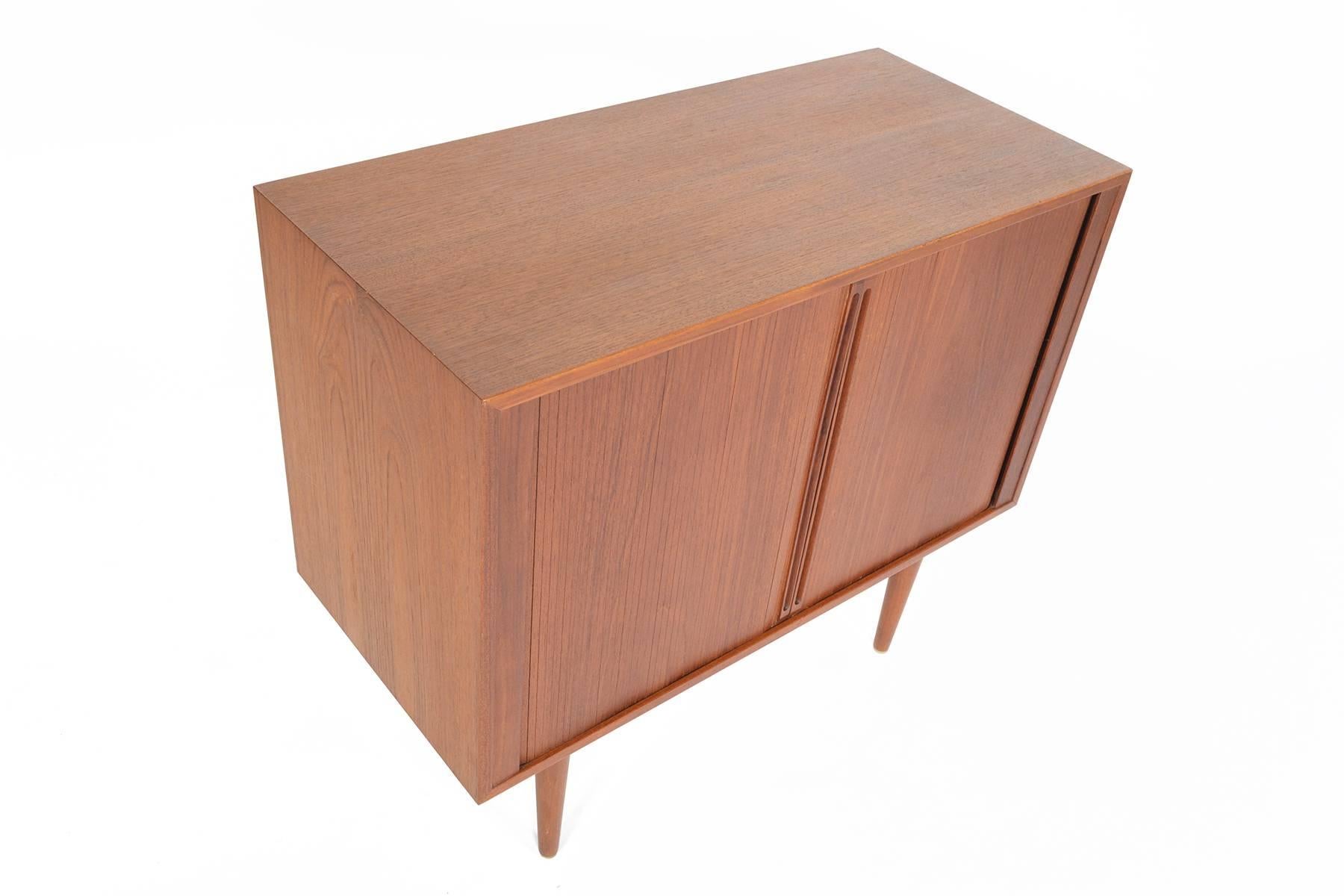 This Danish modern Mid-Century small tambour door credenza in teak was designed by Kai Kristiansen in the 1960s.This fantastic piece features two tambour doors which open to reveal open storage with adjustable shelves and two drawers. In excellent