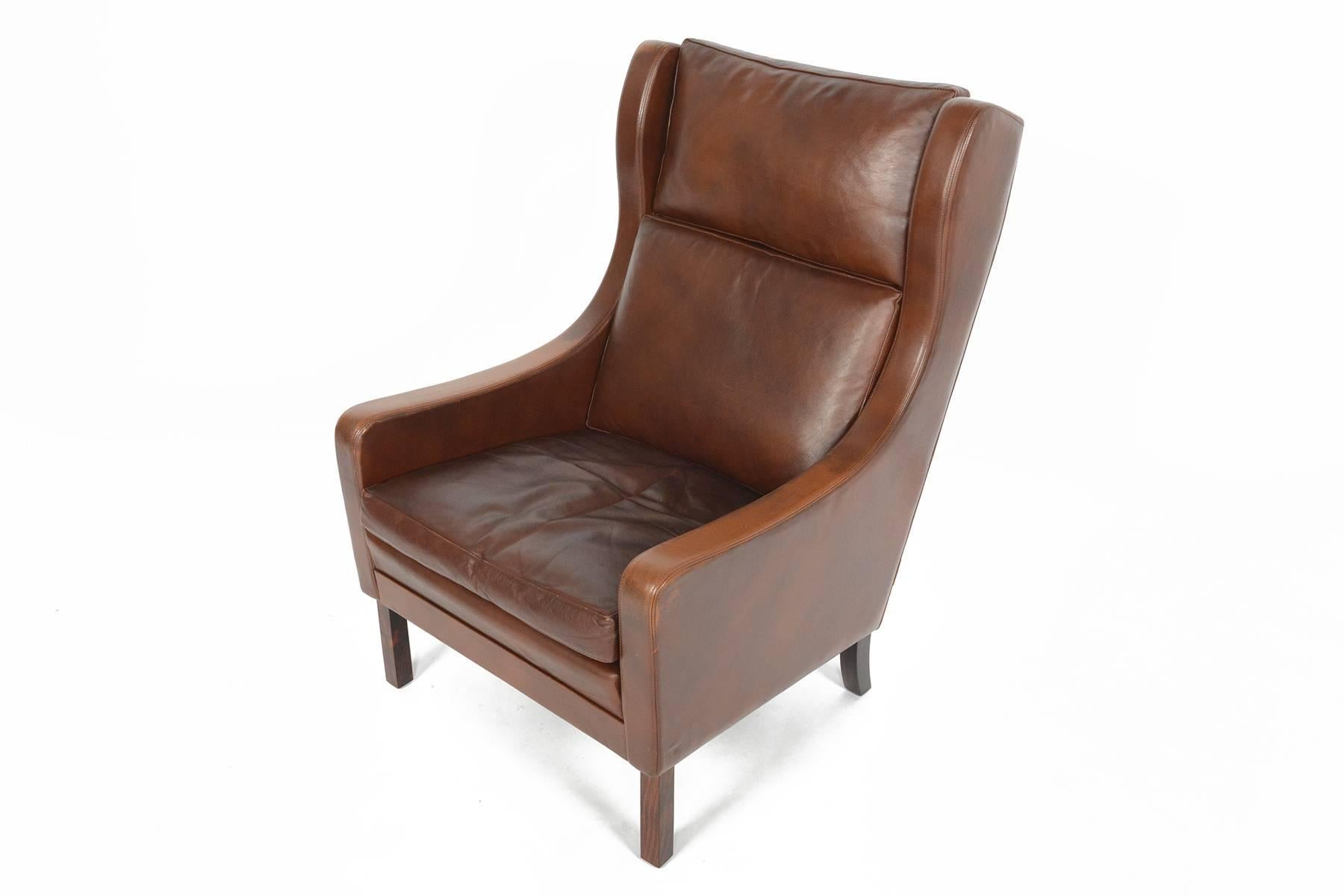 This beautiful Mogensen style high back lounge chair is covered in a rich patinated brown leather. Three leather cushions rest in the winged frame. Chair stands on four tapered Brazilian rosewood legs. In excellent condition with typical wear for
