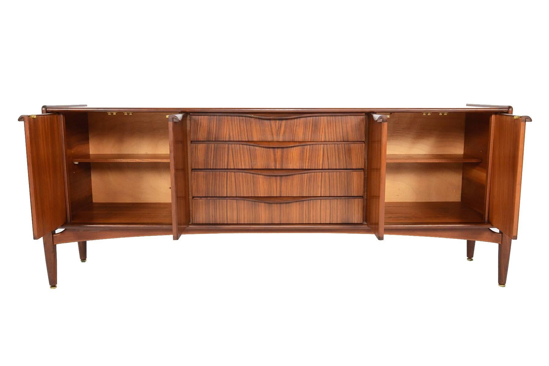 This English modern Mid-Century credenza in mahogany and tola was manufactured by Elliott's of Newbury in the 1960s. This beautiful piece features a floating base, supported by exterior legs with delicate brass hardware. Either end of this sideboard