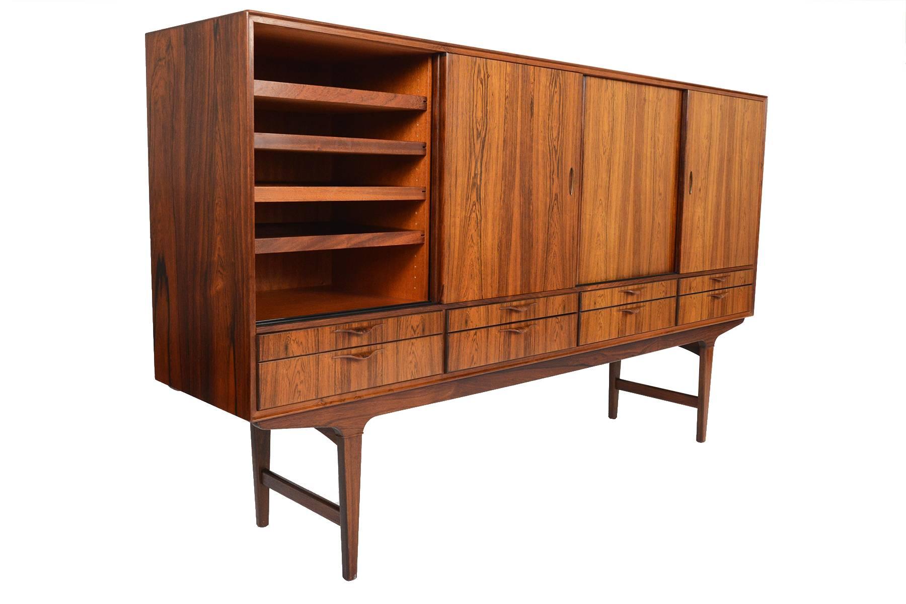 This tall Danish modern credenza is crafted in Brazilian rosewood and showcases gorgeous active grain patterning throughout. Four large doors open to reveal three bays. The left bay houses four adjustable drawers while the centre and right bays