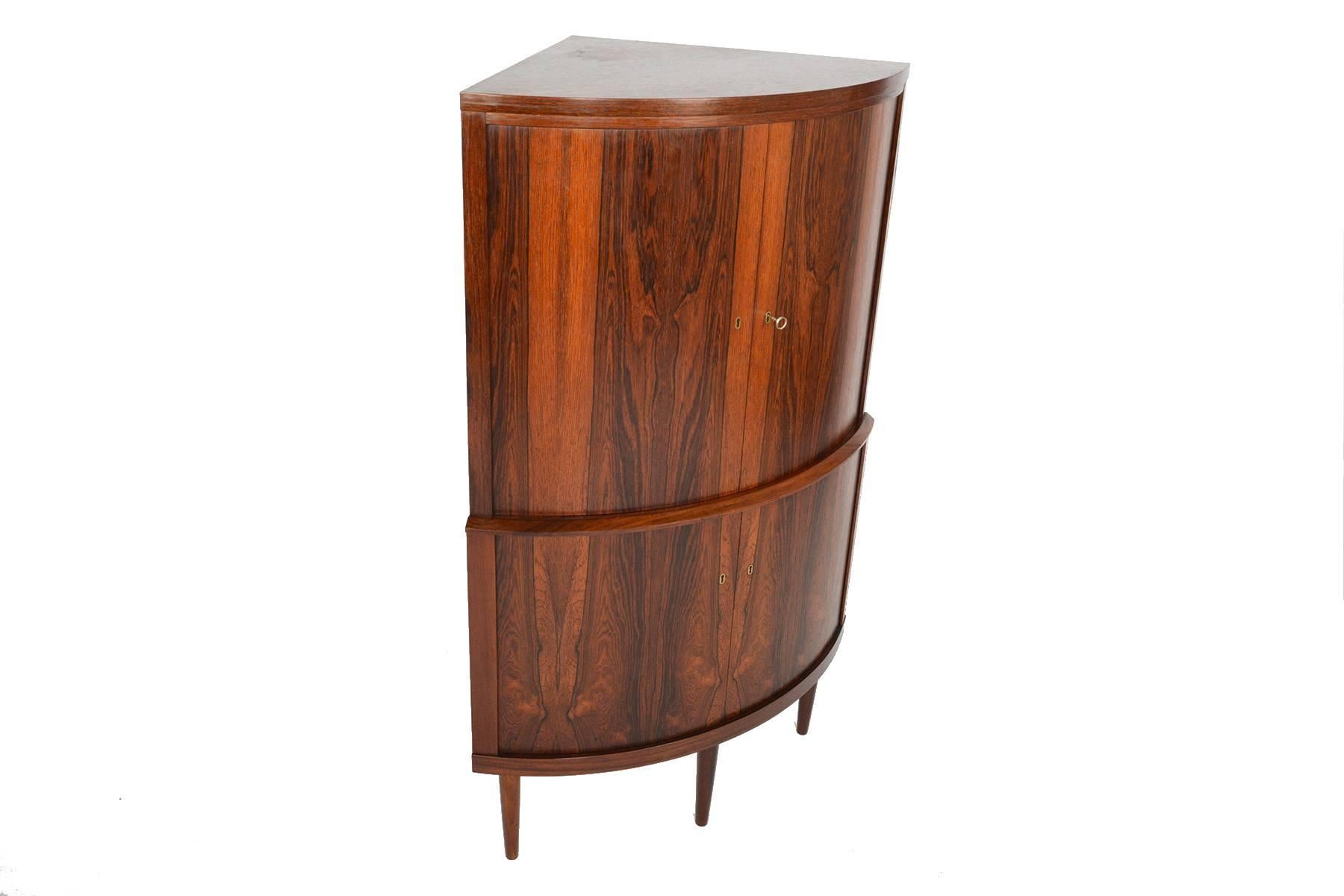 This beautifully understated bow front corner unit in Brazilian rosewood features a Classic and stunning Silhouette. The curved doors unlock onto a large shelved interior. The lower compartment provides additional storage. With a total of three