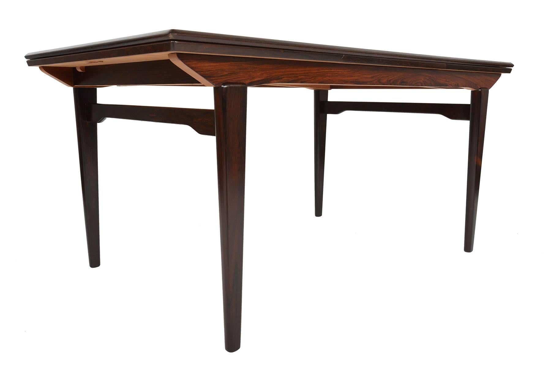 This gorgeous Danish modern Brazilian rosewood dining table offers an angular banding and a well- built braced leg design. Two draw leaves pull-out to almost double the size of the table for entertaining. Recently refinished and in excellent