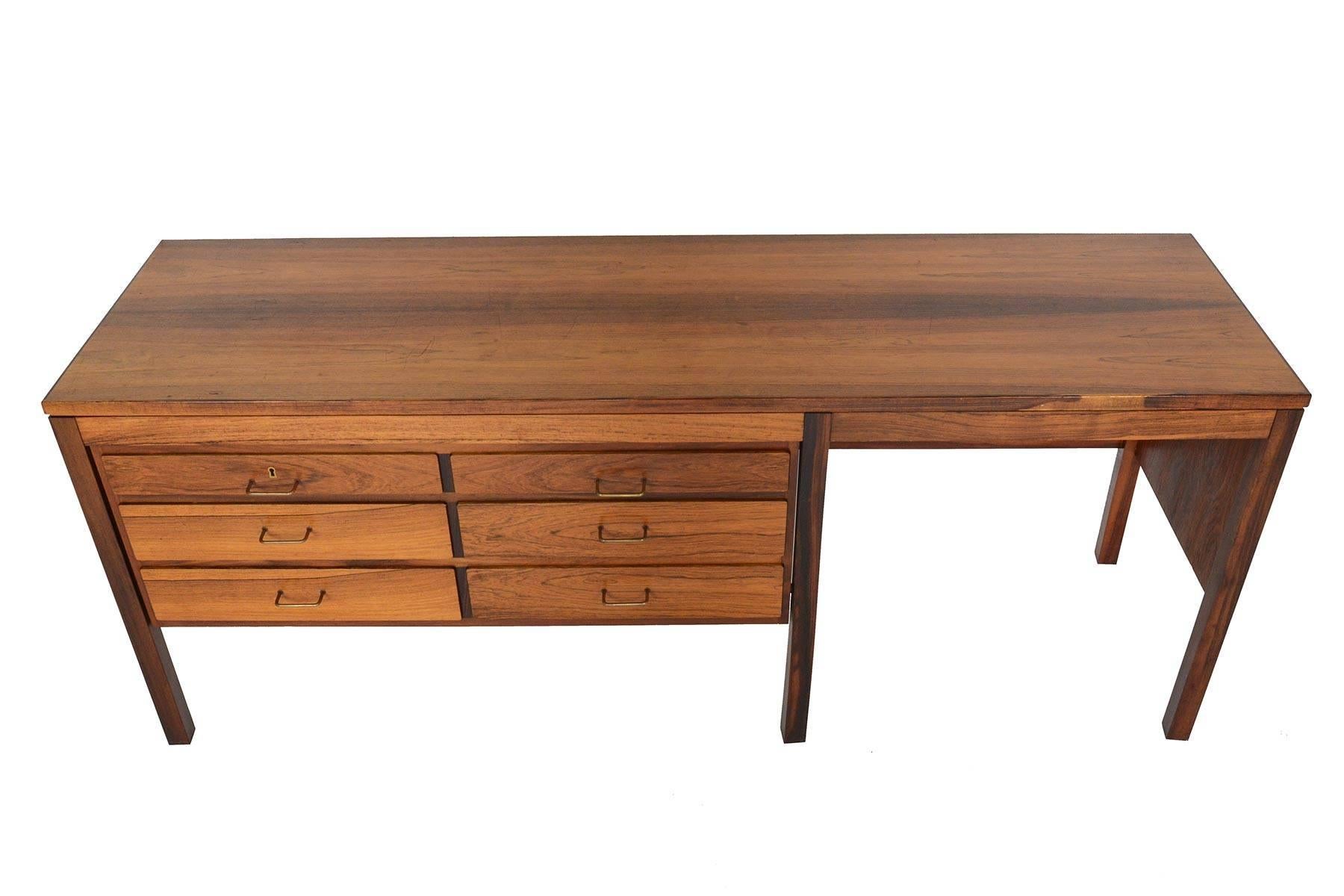 This unusually configured Danish modern dresser vanity is crafted in Brazilian rosewood and provides six large drawers for storage. Each drawer is adorned with original brass pull. An open work surface with wide kneehole sits on the right of this