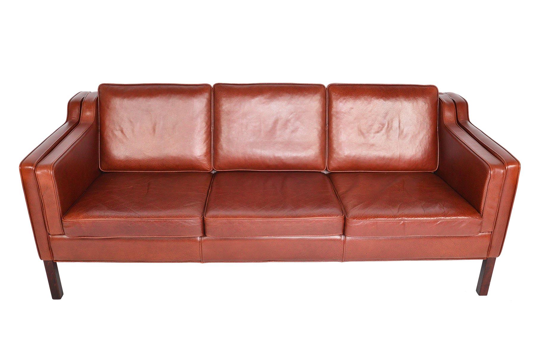 This Danish modern midcentury three-seat sofa in rust red leather by Stouby is a staple of modern decor. This completely original sofa is covered in beautifully grained leather and stands on gorgeous squared mahogany legs. Upholstered on all sides,