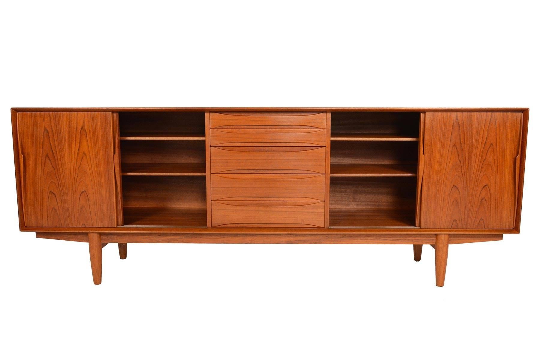 This gorgeous Danish modern midcentury credenza was manufactured by Dyrlund in the 1960s. Beautifully sculpted teak door pulls flank two large doors on each side, which slide open to reveal adjustable shelving. Five centre drawers accented by