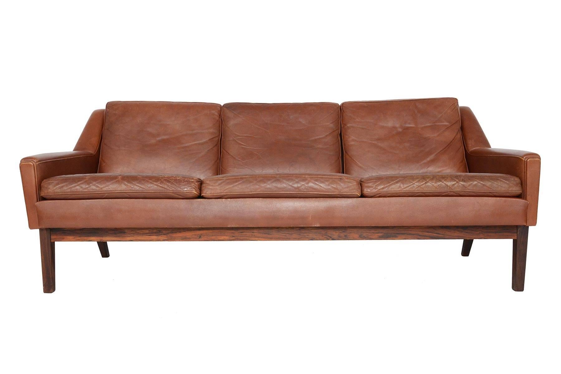 This Danish modern mid century three seat sofa in patinated rust toned leather will make a statement in any modern home. Covered in original leather and raised on a beautiful sculpted rosewood base, this sofa is upholstered on all sides and can be