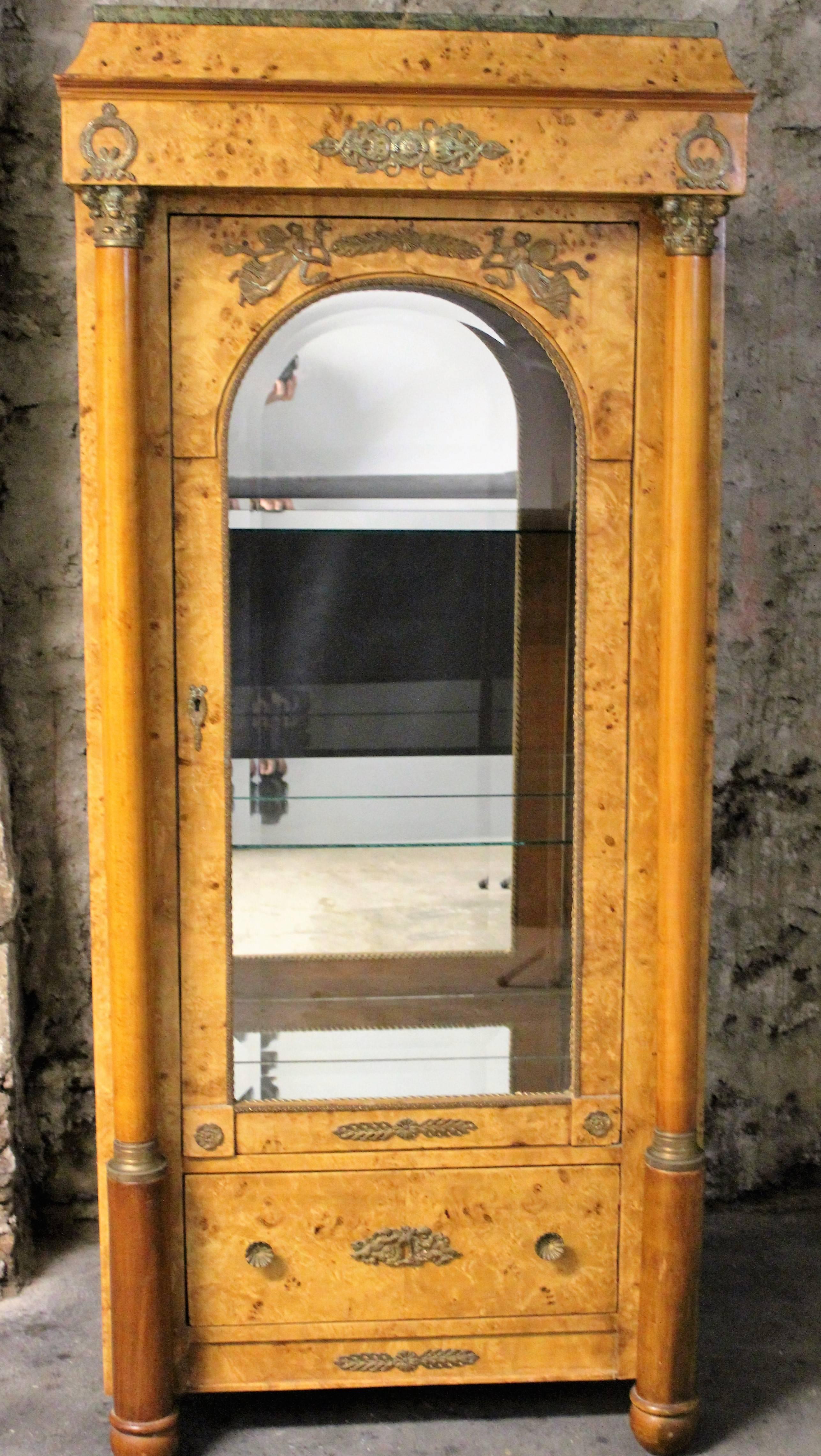This is a stunning French Empire style burl wood vitrine with beautiful ormolu mounts, beveled glass and a marble top. The glazed domed shaped door is flanked with an imposing pair of columns that are capped with exquisite ormolu mounts. The