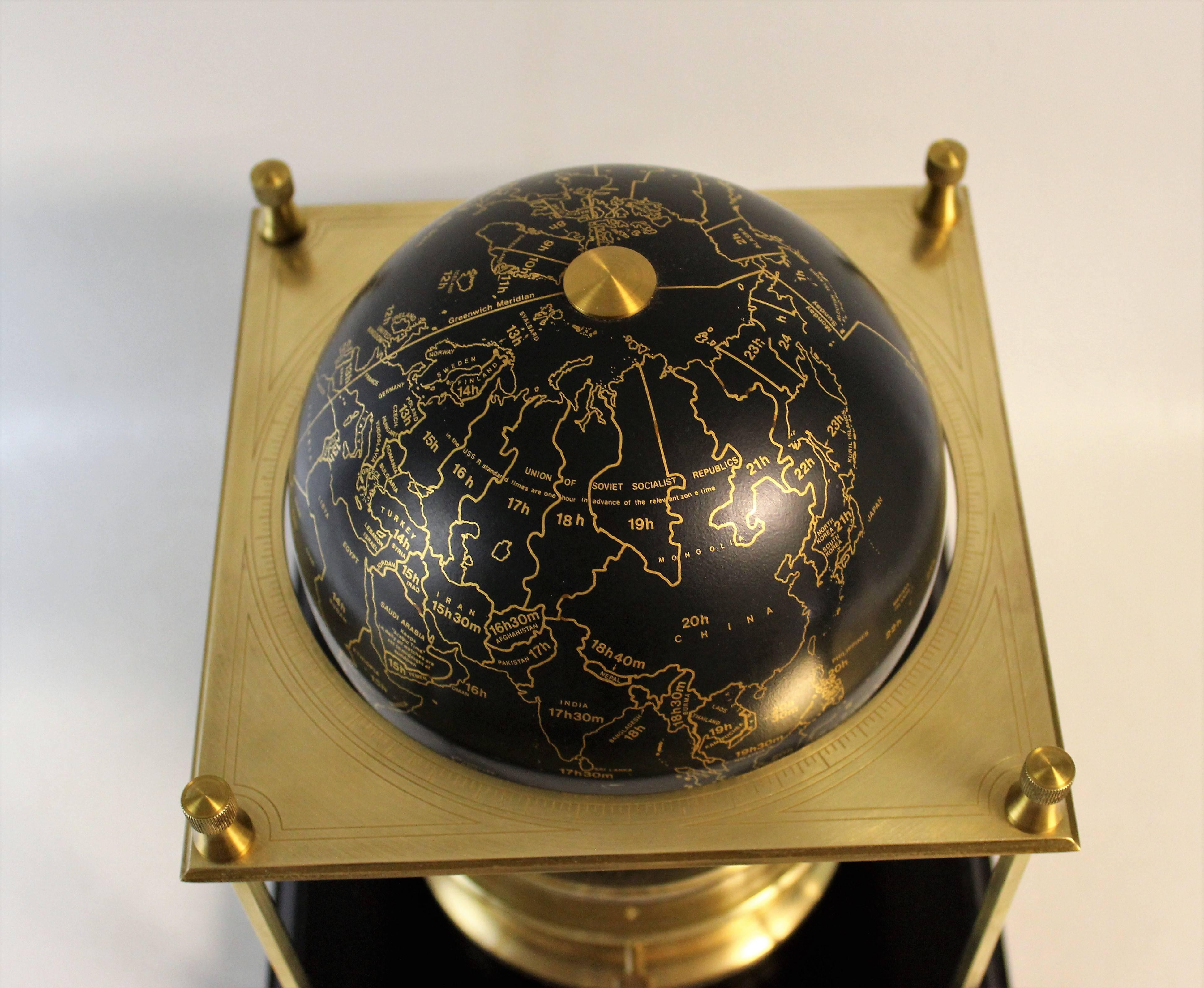 This Royal Geographical society world clock contains a 15 jewel movement by Arthur Imhof S.A. of La Chaux de Fonds Switzerland. The brass square frame with columns supports a terrestrial globe with time zones all marked in gilt on a black metallic