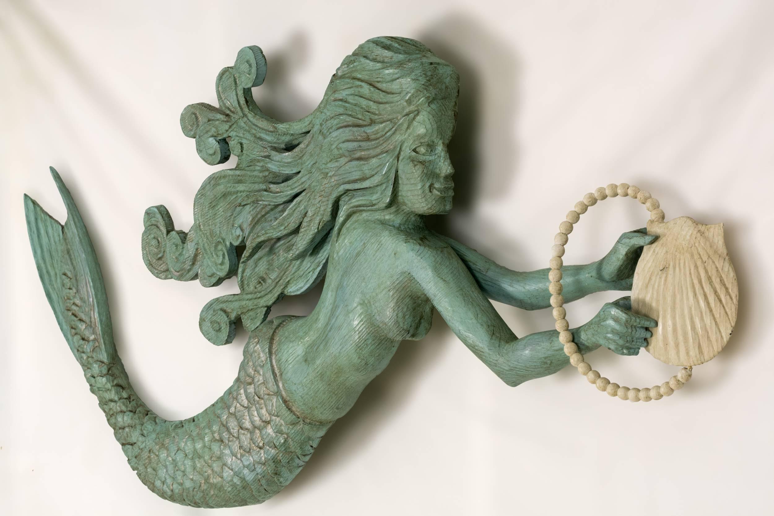 Carved and painted windswept mermaid holding a shell with a string of pearls.
Carved from pine with a verdigris paint finish. Wall-mounted on a metal bracket for three dimensional effect. Carved by American Folk Artist's Jac and