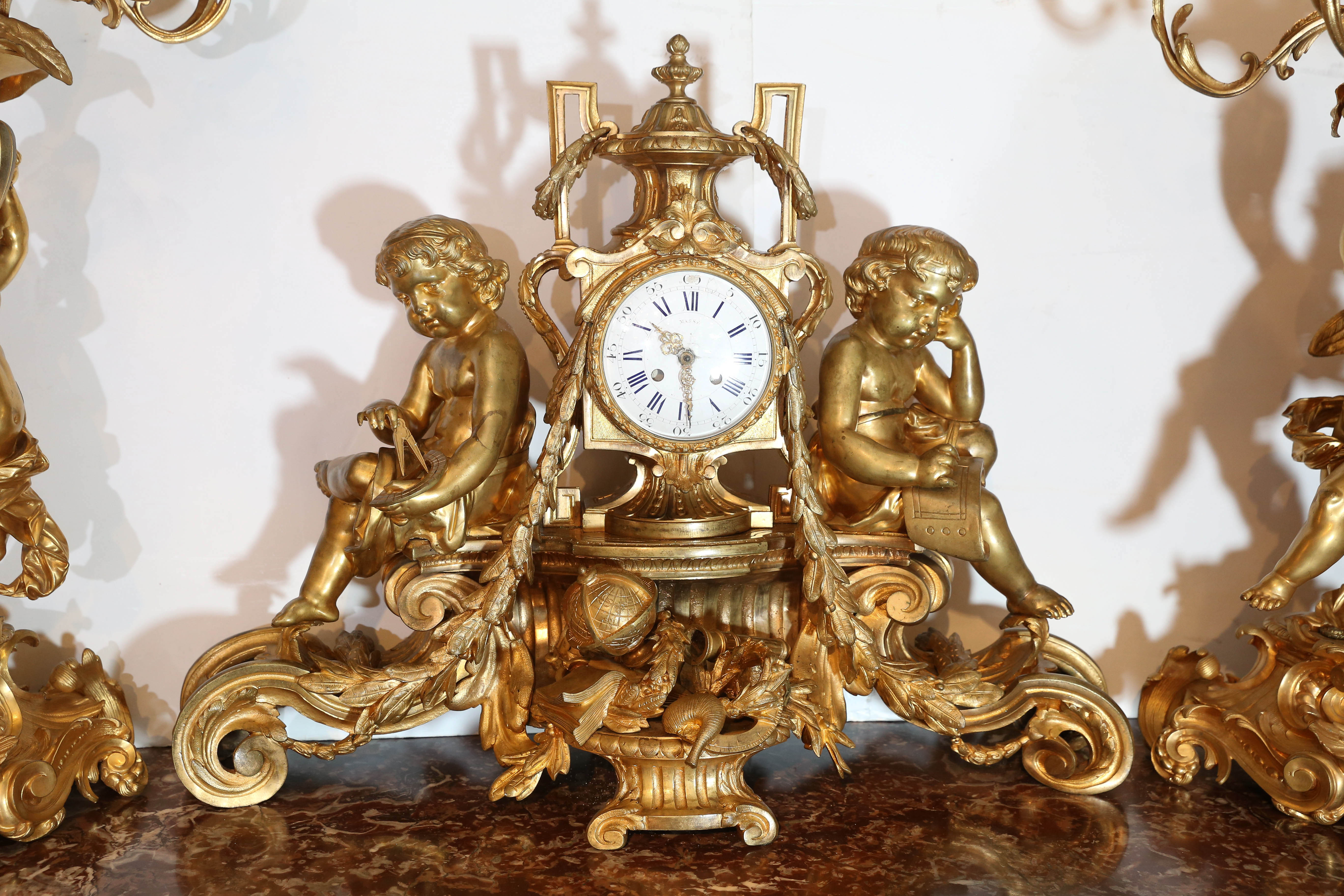 Large three-piece garniture set. Bronze doré clock has enamel face with Masse, Paris on the front. The figures of putti on the sides of the clock are holding a book and astrological instruments. At the base a globe, book and telescope embellish the