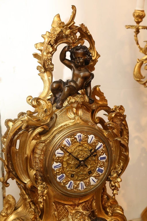 L'orologio in bronzo dorato e il candelabro sono decorati con cherubini in una tonalità scura.
Bronzo patinato. Questo grande ed elaborato set è in ottime condizioni.
Il rococò  Il design incorpora cinque candelabri sul ramo.
Tenuto da un