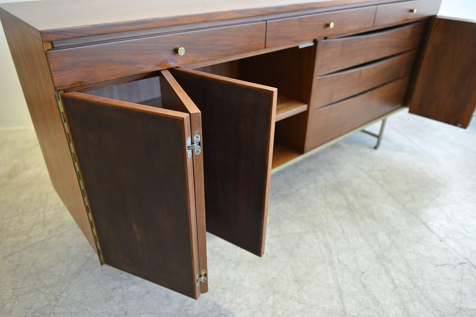 Rare Paul McCobb for Calvin Group walnut and leather front credenza with Classic flat bar brass legs. Accordion trifold doors open to reveal adjustable shelving on left and three drawers on the right. Upper smaller drawers are all felt lined for
