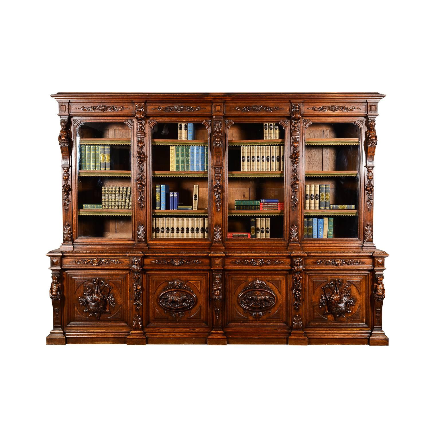 A monumental eight-door French ‘Hunters’ library bookcase in solid oak, with the lower doors featuring hand-carved panels of fox, hunting dog, and game-birds with hunting accessories. The upright pilasters are embellished with cherubs and swags of