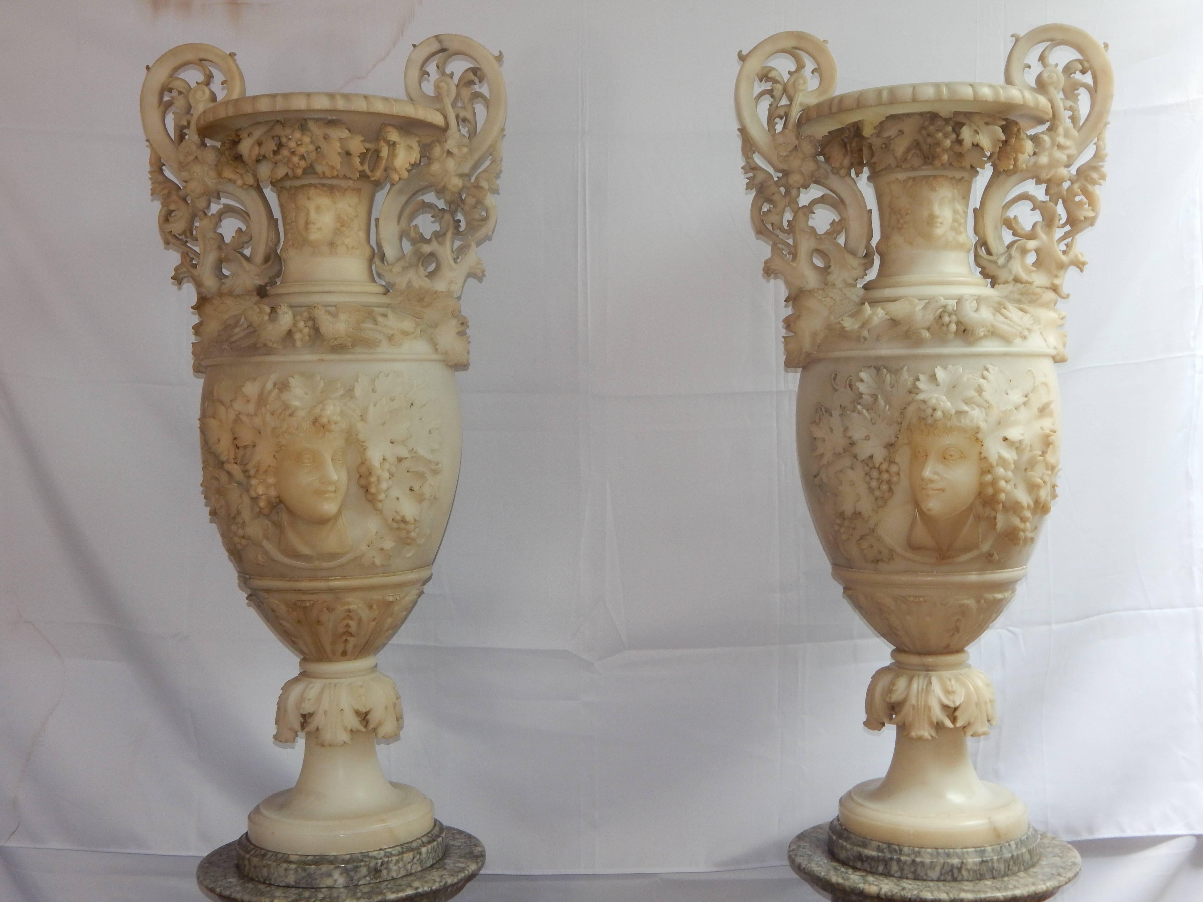 An incredible pair of monumental carved alabaster urns with pedestals.
The double handled urns scroll down to reveal a Griffin's head, which sits above a bacchante mask. Dripping with grapes and leaves throughout, this pair of seven and a half foot