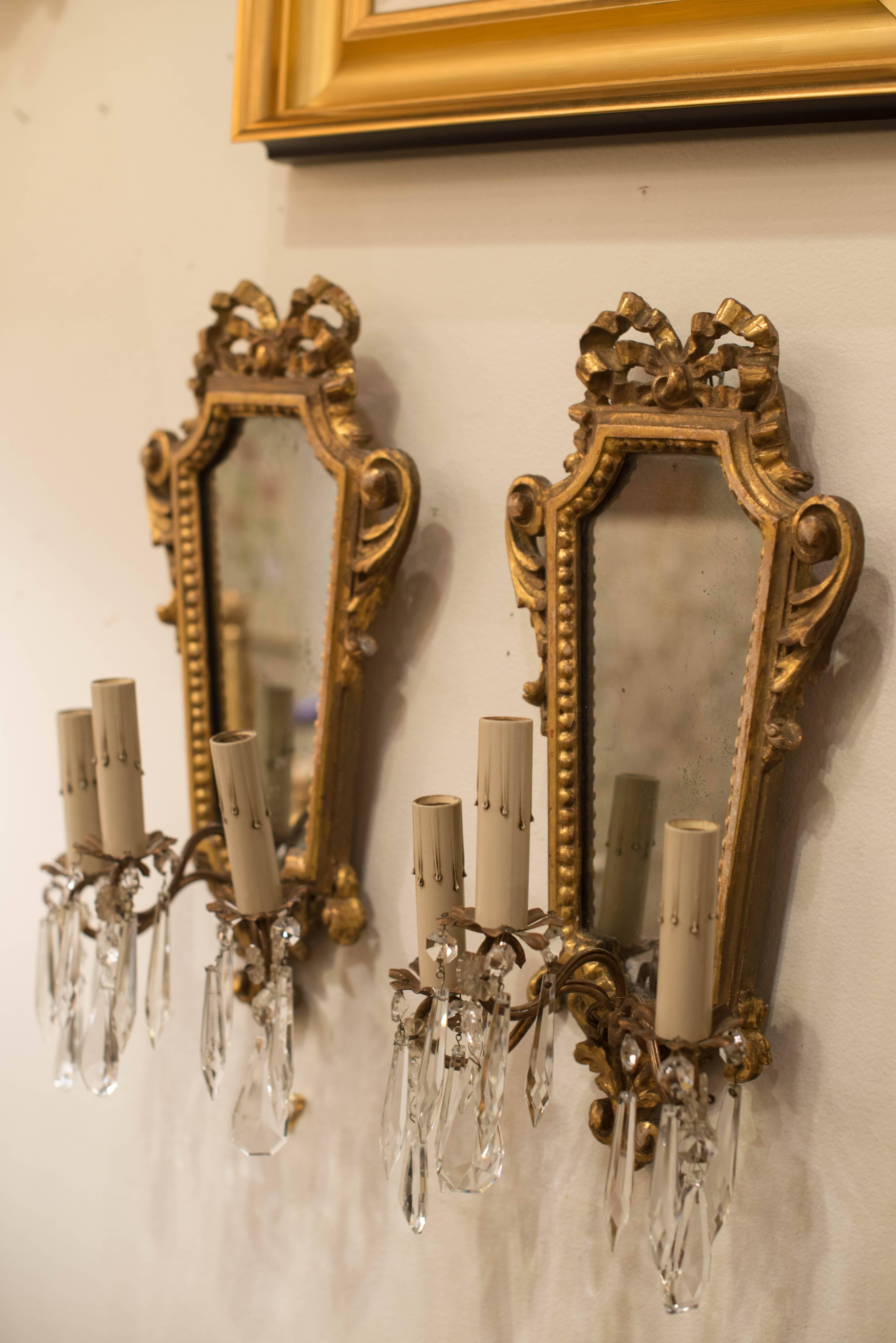 This pair of 19th Century Italian giltwood three arm sconces features a mirrored back and a lovely bow and acanthus motif.  The arms have crystal prisms in different shapes and sizes.  These sconces have been newly rewired.